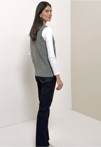 Gilet sans manches en maille grise porté sur une chemise blanche à manches longues, associé à un jean bleu foncé. Texture lisse avec une coupe ajustée.