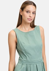 Hellgrünes, ärmelloses Kleid mit rundem Ausschnitt, tailliertem Oberteil und plissiertem Rock. Hergestellt aus glattem Stoff mit dezentem Glanz.