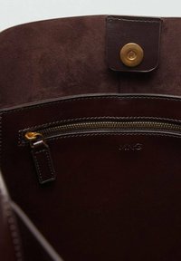 Dark brown leather bag interior with a gold zipper pocket and a magnetic snap closure on the flap, embossed with "MNG" logo.