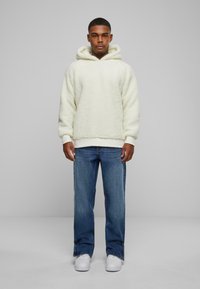Cream-colored fleece hoodie with a kangaroo pocket, ribbed cuffs, and hem; paired with blue straight-leg jeans and white sneakers.
