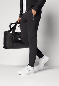 Un pantalon de sport noir avec un petit logo, associé à des baskets blanches ; un sac de sport noir est tenu par une sangle.