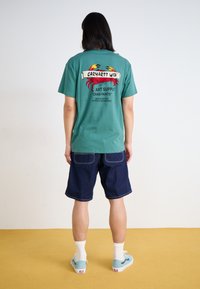 Camiseta de algodón color azul verdoso con estampado gráfico de un cangrejo y loros, combinada con pantalones cortos de mezclilla azul oscuro y zapatillas de deporte azul claro. Calcetines blancos visibles.