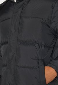 Blouson matelassé noir avec capuche, présentant un design matelassé, des poches latérales et une finition texturée brillante. Petit logo sur la poitrine.