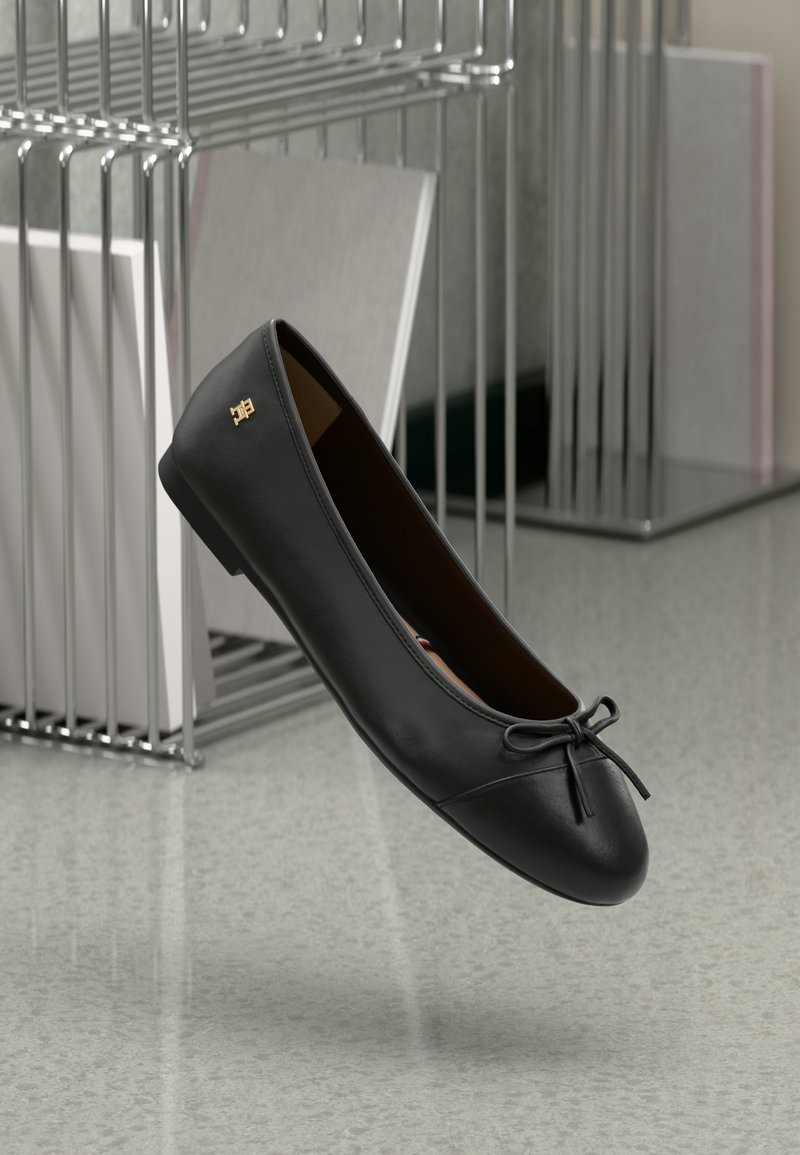 Ballerines en cuir noir avec un petit nœud à l'avant et un logo doré sur le côté, positionnées près d'étagères en métal sur un sol à motifs.