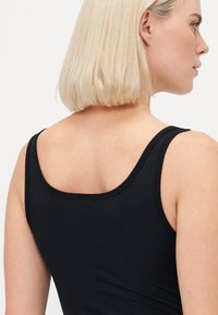 Žena s krátkými blond vlasy, oblečená v černém topu bez rukávů, zobrazena zezadu a částečně v profilu na jednobarevném pozadí.