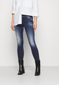 Jeans de denim ajustados en azul oscuro con acentos descoloridos, combinados con una camisa blanca de botones y botas de tacón negras, creando un look casual.