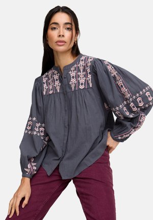 Frau trägt eine lockere dunkelgraue Bluse mit pinker geometrischer Stickerei an Ärmeln und Brust, kombiniert mit dunkelroter Hose, posiert vor einem neutralen Hintergrund.