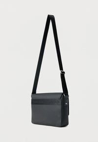 Μαύρη ορθογώνια τσάντα χιαστί με ρυθμιζόμενο λουράκι, ανάγλυφο λογότυπο "Lacoste" στο καπάκι και το λουράκι, μικρό έμβλημα κροκόδειλου στο πλάι.