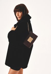 Cappotto in pelliccia sintetica nera con colletto alto, abbinato a una borsa a tracolla in cuoio marrone scuro con finiture goffrate, dotata di una placca con logo dorato e tracolla a catena.