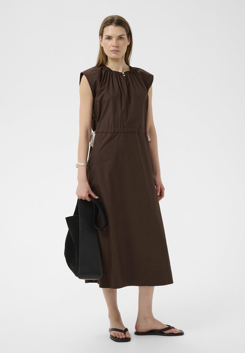 Femme portant une robe midi sans manches marron foncé, tenant un grand sac cabas noir, debout avec des tongs noires sur un fond uni.