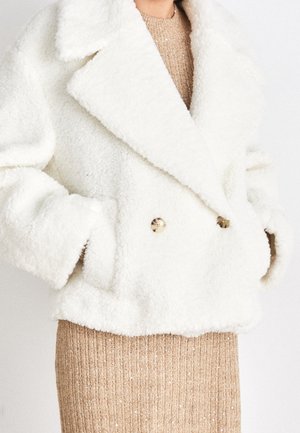 Person trägt einen weißen, flauschigen, zweireihigen Mantel mit goldenen Knöpfen über einem beige gerippten Kleid mit dezenten Glitzereffekten.