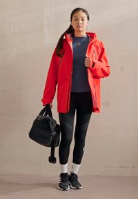 Red waterproof jacket, blue fitted shirt, black leggings, black sneakers, and a black bag. The jacket has a hood and zippered pockets.