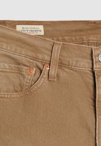 Jean en denim beige présentant un design standard à cinq poches, un bouton métallique et des accents cuivrés. L'étiquette indique "Levi's Premium."