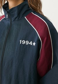 Námořnická větrovka s maroon ramenními panely, bílými detaily, zapínáním na zip a vyšitým "1994" se hvězdou na přední straně.