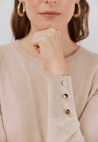 Beige Strickpullover mit gerippten Bündchen, ausgestattet mit drei dekorativen Knöpfen: zwei metallisch und einer mit einem schwarzen Steindetail. Weicher Stoff erkennbar.