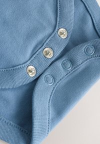 Tissu bleu avec boutons-pression assortis et boutons métalliques argentés sur un bord courbé, montrant les détails de la fermeture du vêtement.