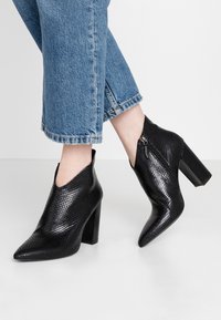 Schwarze, hochhackige Stiefeletten aus strukturiertem Kunstschlangenleder. Mit einer spitzen Zehenpartie und einem seitlichen Reißverschluss. Vor einem Hintergrund aus blauen Jeans.
