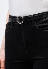 Κοντινή λήψη ενός ατόμου που φοράει μαύρο τζιν με μαύρη δερμάτινη ζώνη που έχει ασημένια στρογγυλή αγκράφα και λευκό πουκάμισο βαλμένο μέσα.