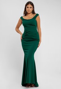 Vrouw in een aansluitende, donkergroene, off-shoulder avondjurk, staande met één hand op haar heup tegen een effen achtergrond.