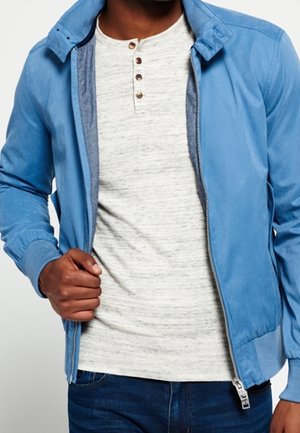 Mann trägt eine hellblaue Bomberjacke über einem hellgrauen Henley-Shirt mit Holzknöpfen, kombiniert mit dunkelblauen Jeans.