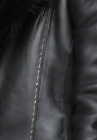 Schwarze Lederjacke mit Reißverschluss, glatter Textur und einem Pelzkragen. Weiches Leder mit sichtbaren Nähten und metallischen Reißverschluss-Akzenten.