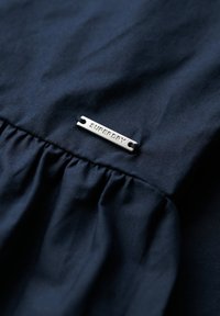 Granatowy materiał z plisowanym detalem; na metce znajduje się srebrny metalowy tag z wytłoczonym napisem "SUPERDRY". Gładka tekstura, casualowy design.