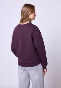 Fioletowa bluza z okrągłym dekoltem i ściągaczami, charakteryzująca się luźnym krojem. Noszona z jasnoszarymi dżinsami, prezentując prosty design.