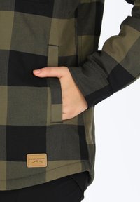 Groen en zwart geruit flanellen shirt met een borstzak en zijhandzak; heeft een leren label met het "Swedemount" logo aan de onderkant.