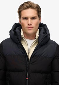 Czarna kurtka puchowa z kapturem, o lekkiej konstrukcji, z zapięciem na zamek i kontrastowymi teksturowanymi panelami. Udekorowana metką z logo.