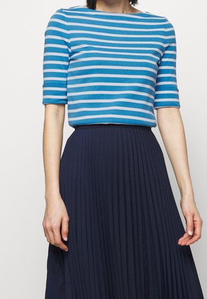 Vrouw die een blauw-wit gestreept kortärmelig shirt draagt, ingestopt in een hooggetailleerde, marineblauwe geplooide rok, zichtbaar van nek tot bovenbenen.
