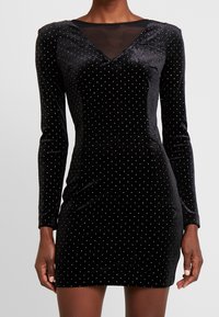 Schwarzes Samtkleid mit langen Ärmeln, V-Ausschnitt und transparenter Netz-Einlage. Verziert mit kleinen goldenen Punkten und hat eine körpernahe Silhouette.