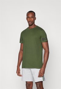 Grønn bomulls T-skjorte med rund hals og korte ermer, med en svart logo på venstre erme. Kombinert med grå shorts.
