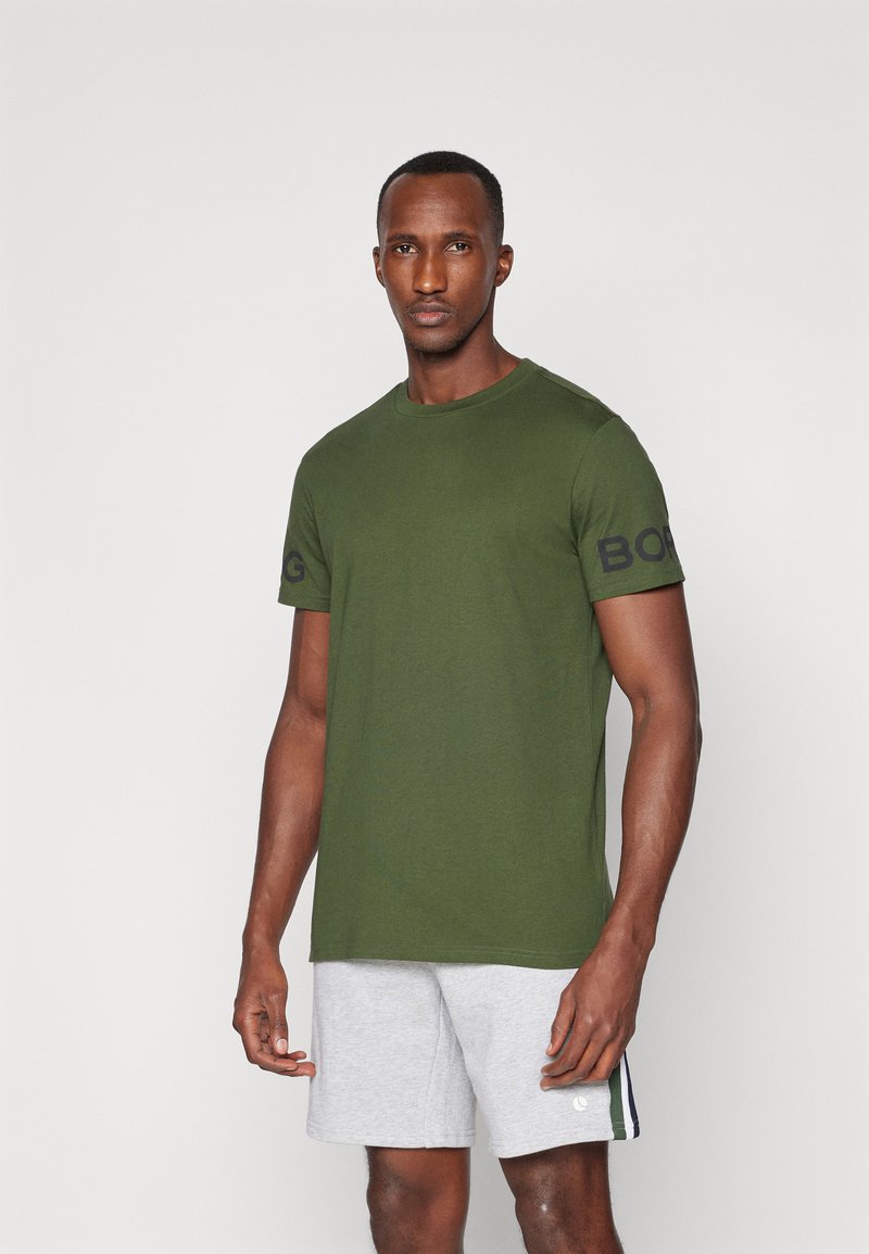 Grønn bomulls T-skjorte med rund hals og korte ermer, med en svart logo på venstre erme. Kombinert med grå shorts.