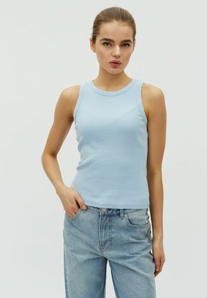 Jauna sieviete, tērpusies gaiši zilā bezroku ribotā topā un vaļīgās gaiši zilās džinsās, pozē uz vienkārša baltā fona.