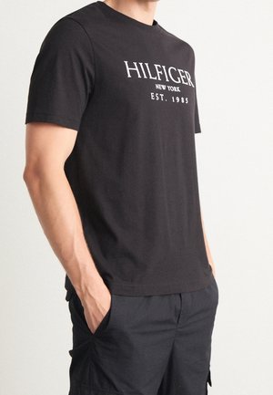 Mann trägt schwarzes Kurzarm-T-Shirt mit weißem Text "HILFIGER NEW YORK EST. 1985" und schwarze Hose, Hände in den Taschen, neutraler Hintergrund.