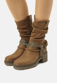 Botas de tobillo de ante marrón con parte superior arrugada, detalles trenzados y una correa decorativa con un patrón floral. Suela robusta y texturizada.