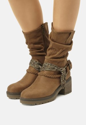 Botas de tobillo de ante marrón con parte superior arrugada, detalles trenzados y una correa decorativa con un patrón floral. Suela robusta y texturizada.