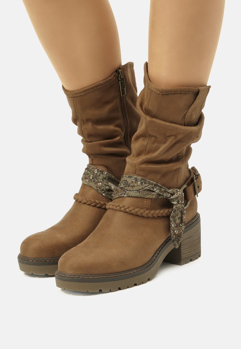 Botas de tobillo de ante marrón con parte superior arrugada, detalles trenzados y una correa decorativa con un patrón floral. Suela robusta y texturizada.