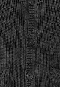 Juodas megztas kardiganas su tekstūruotu ribbingo raštu, didelėmis juodomis sagomis ir šoninėmis kišenėmis. Artimas vaizdas į priekines detales.