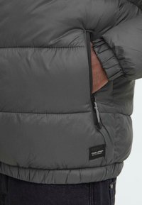 Grå pufferjakke med en quiltet design, der har en sidelukning og elastiske manchetter, lavet af letvægts, vandafvisende materiale.