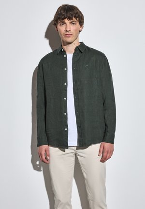 Ciemnozielona lniana koszula z guzikami i kołnierzykiem, noszona na białym T-shircie, w połączeniu z jasnoszarymi spodniami. Prosty design, bez wzorów.