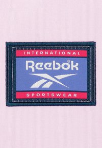 Parche con el logo de Reebok sobre un fondo azul, texto y símbolo en blanco, borde negro, sobre tela rosa. El texto dice "International Sportswear".