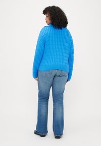 Pulover tricotat albastru cu un model texturat, asortat cu blugi evazați de culoare albastru deschis, văzut din spate.