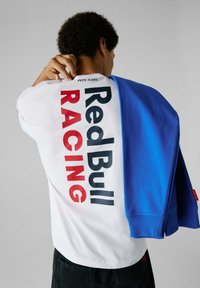 Camiseta blanca con gran texto "Red Bull RACING" en rojo y azul oscuro. El modelo lleva un sudadera azul colgada sobre un hombro. Jeans negros.