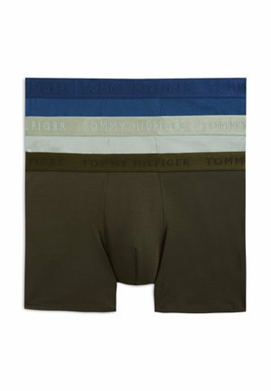 Młody mężczyzna stoi boso, ubrany w oliwkowozielone bokserki, zwrócony przodem na jednolitym białym tle.