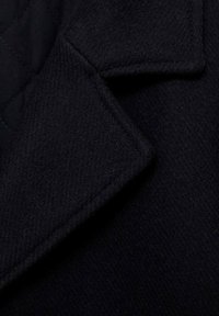 Svart ullrockstyg med ett vadderat innerfoder. Har en strukturerad krage, fin textur och en ren, skräddarsydd look.