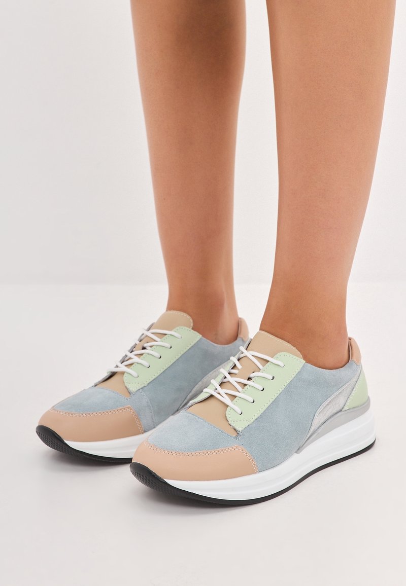 Jambes portant des baskets aux couleurs pastel avec des panneaux beige, bleu clair et vert menthe et des lacets blancs, debout sur une surface blanche unie.