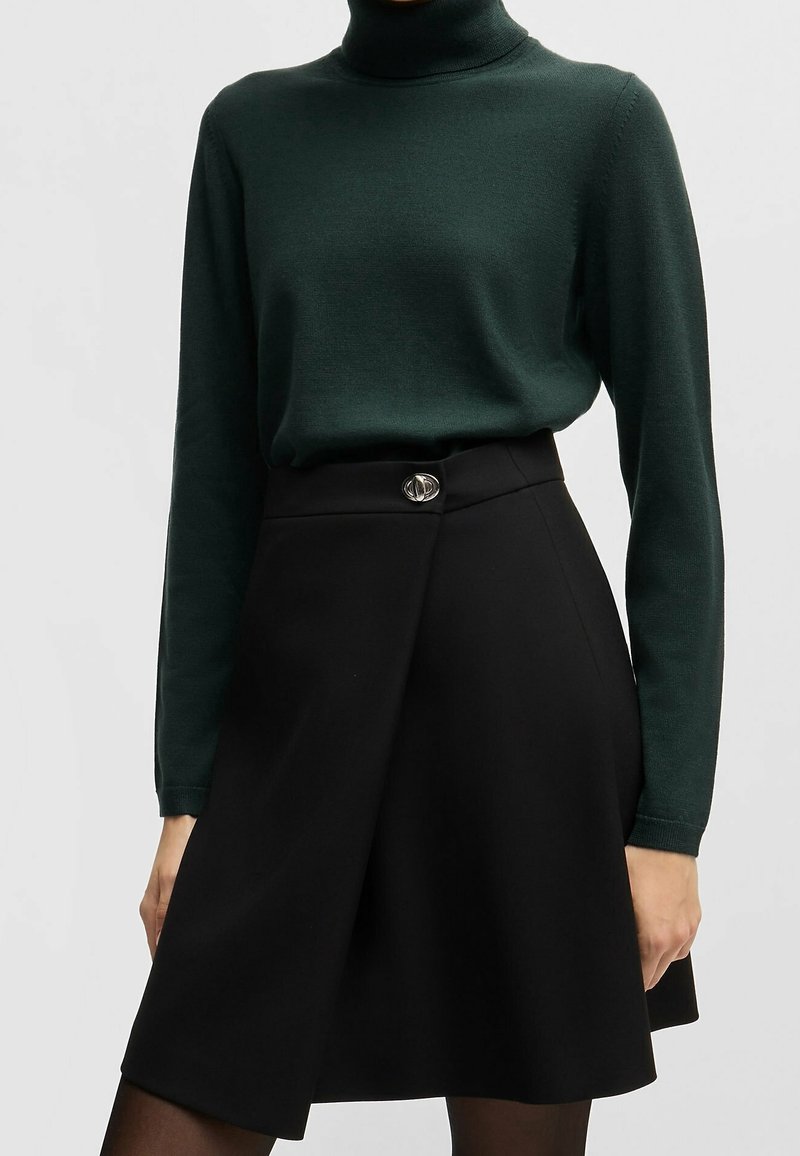 Mörkgrön polotröja kombinerad med en svart A-linjeformad kjol med en silveraccentknapp vid midjan. Slät tygtextur.