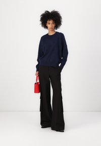 Marineblå strikkegenser med teksturert mønster, vide sorte bukser og en liten rød håndveske. Lange ermer og avslappet passform.