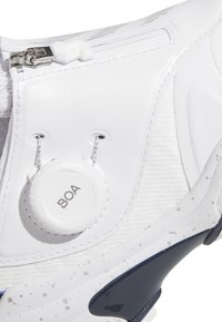 Bílá teniska s texturovaným svrškem, zapínáním na zip, nastavitelným kolečkem BOA a tmavě modrými akcenty na podrážce. Viditelné vzorované švy.